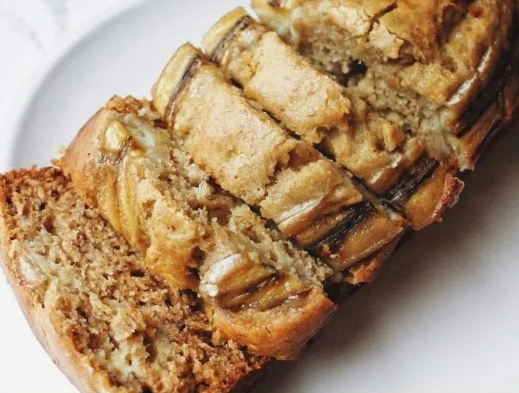 A slice of fluffy banana bread topped with nuts and fresh bananas on a wooden table.